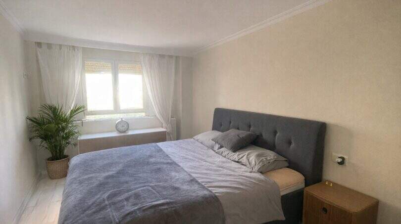 Luminoso dormitorio con cama doble, cabecero tapizado gris, mesilla de noche de madera y planta de interior cerca del mar