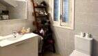 Helles Badezimmer der Mallorca-Finca mit weißem Waschtisch, Holzregal und modernem WC, graue Fliesen und Holzboden.