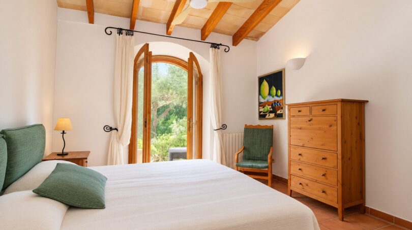 Helles Schlafzimmer der Natursteinfinca mit Holzbalkendecke, Doppelbett und Blick in den Garten.