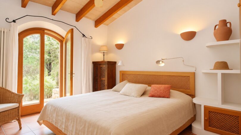 Helles Schlafzimmer der Natursteinfinca mit Holzbalkendecke, Terrakottaboden und Bogentür zum Garten.