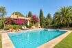 Natursteinfinca mit türkisem Pool, Bougainvillea-Bewuchs, Palmen und mediterranem Garten bei blauem Himmel.