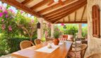 Überdachte Terrasse der Natursteinfinca mit Holzbalken, Esstisch, Rattan-Stühlen und blühenden Bougainvilleas