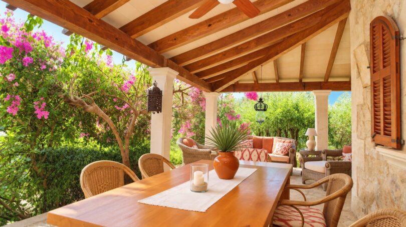 Überdachte Terrasse der Natursteinfinca mit Holzbalken, Esstisch, Rattan-Stühlen und blühenden Bougainvilleas