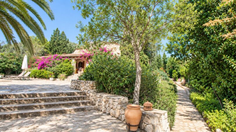 Gepflegter Gartenweg mit Natursteinmauer, Terrakottatopf, Palmen und pinker Bougainvillea vor der Finca.