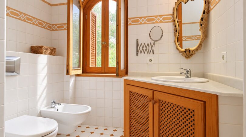 Elegantes Finca-Bad mit Holzschränken, Goldrahmenspiegel, Bidet und dekorativem Fliesenmuster in Weiß-Orange.