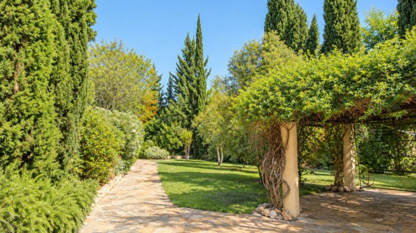 Gepflegter Gartenweg der Natursteinfinca mit Pergola, Zypressen und grüner Rasenfläche.