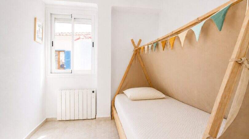 Kinderzimmer mit Tipi-Bett aus Holz, bunten Wimpeln und Heizkörper unter weißem Fenster in Palma-Stadtwohnung
