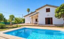 Finca blanca en Palmanyola con piscina azul, detalles de piedra, persianas de madera y vegetación exuberante bajo un cielo despejado.